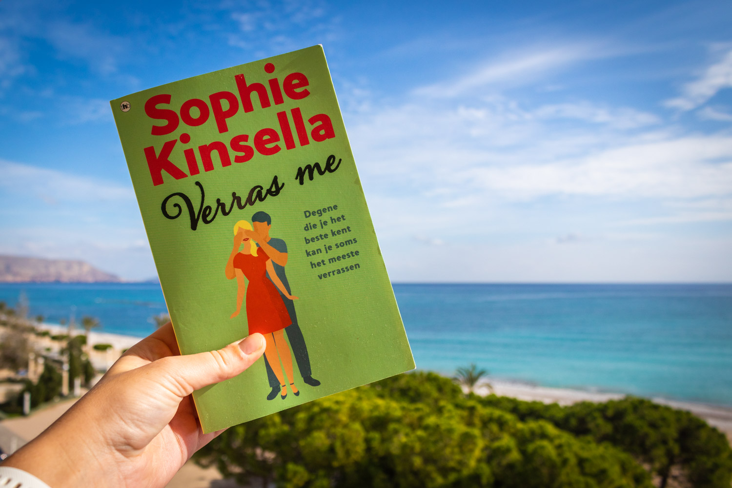 Groene cover van het boek Verras Me van Sophie Kinsella tegen een blauwe lucht en zee.