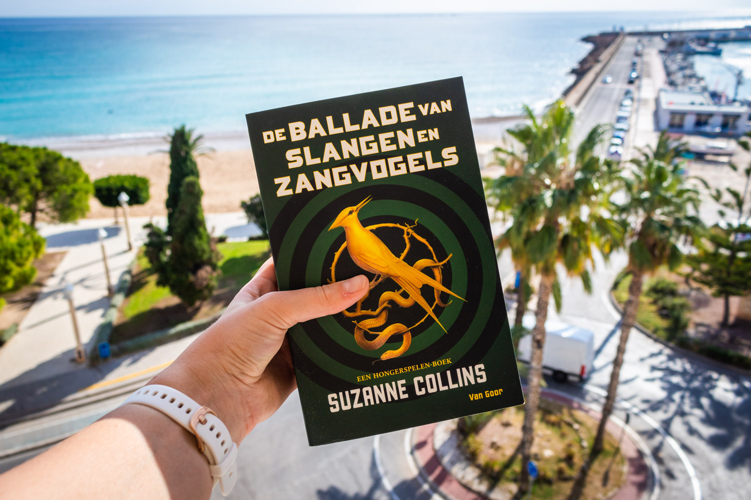 Het boek De ballade van slangen en zangvogels van Suzanne Collins omhoog gehouden met op de achtergrond de Middellandse Zee en palmbomen