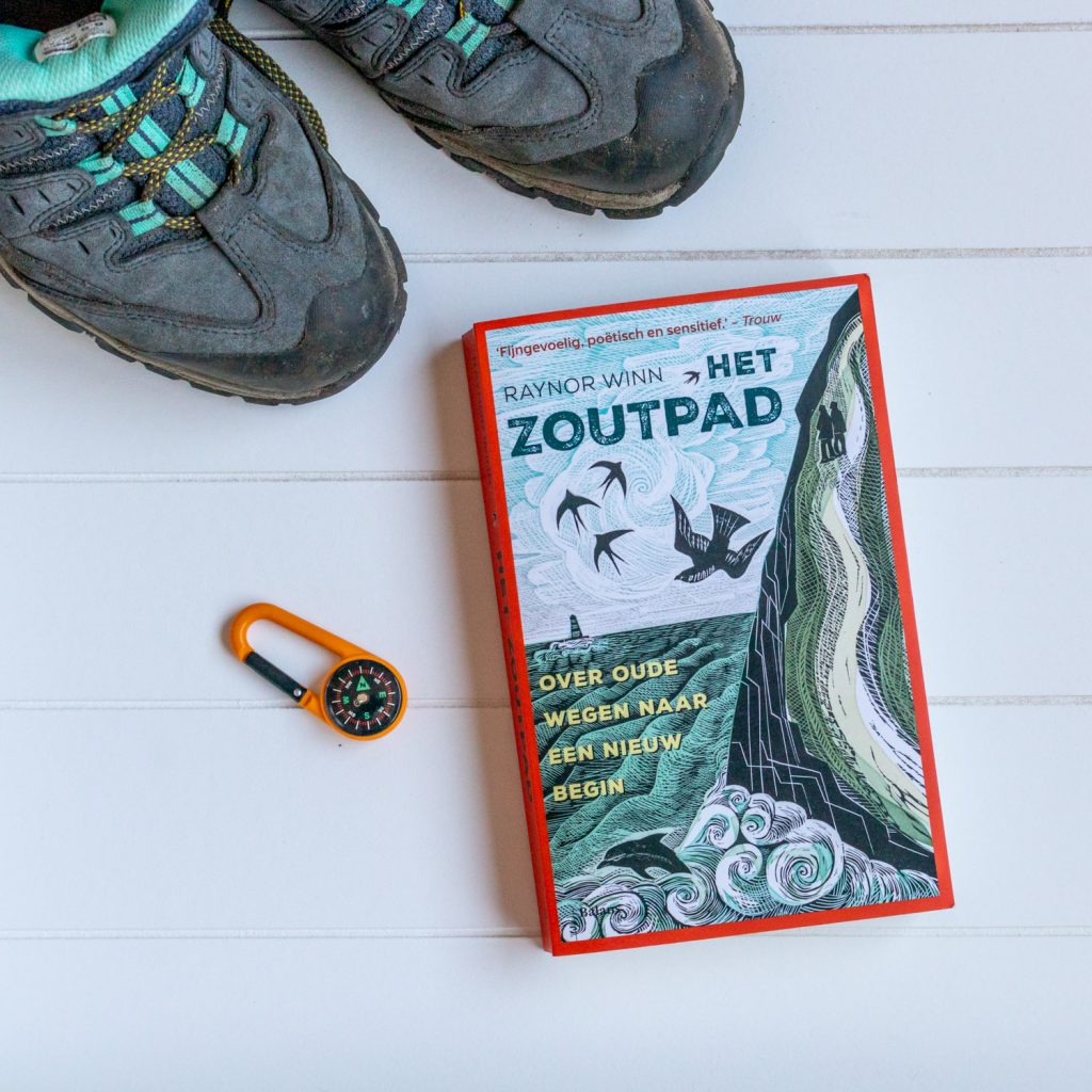 Het boek Het zoutpad van Raynor Winn met een kompas en een paar wandelschoenen