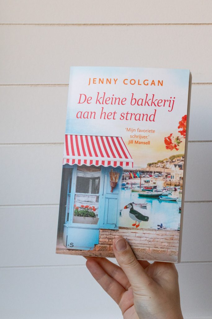 Een hand houdt het boek De kleine bakkerij aan het strand omhoog