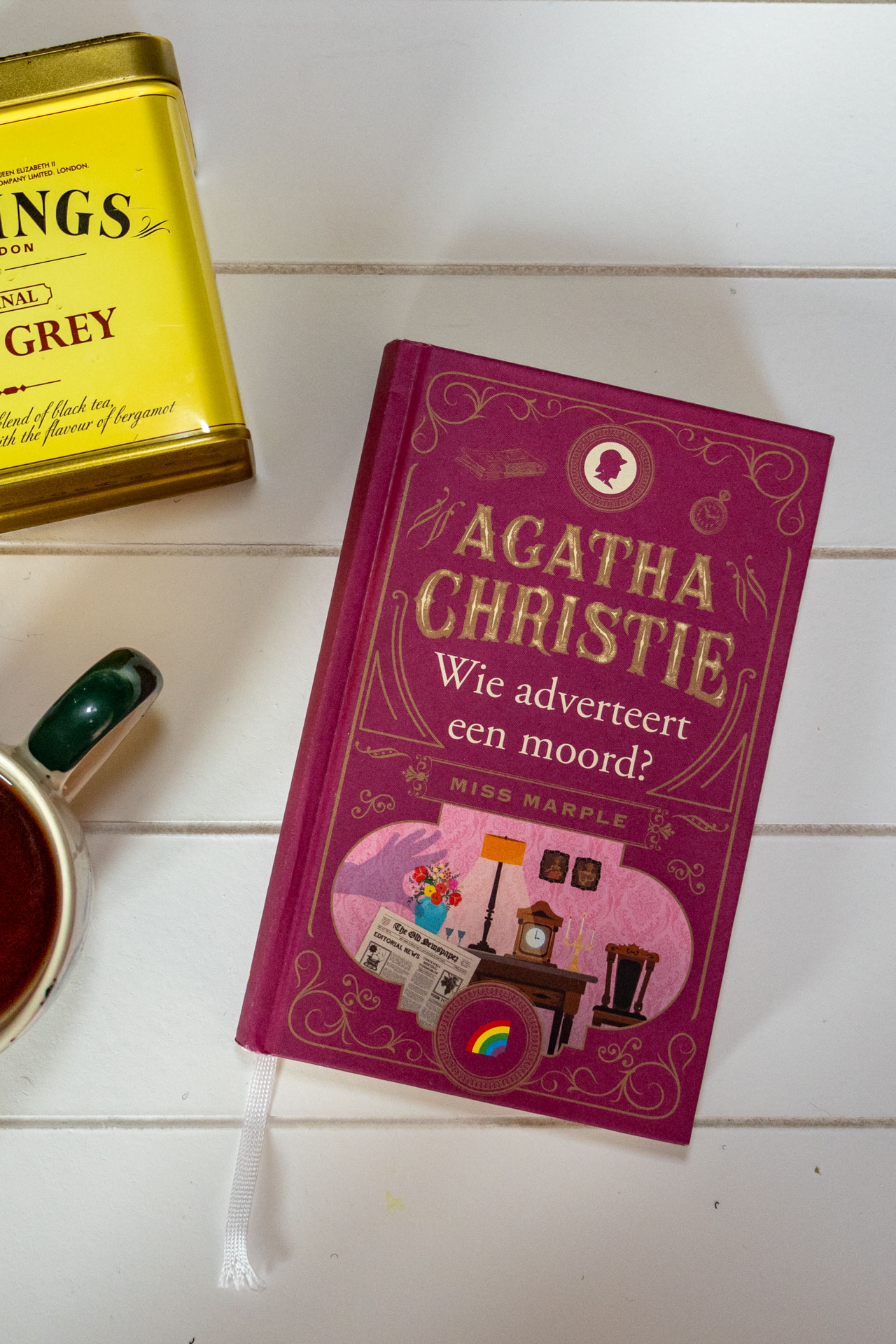 Boek Wie adverteert een moord! van Agatha Christie tegen een witte achtergrond met een kop thee en een blikje losse thee