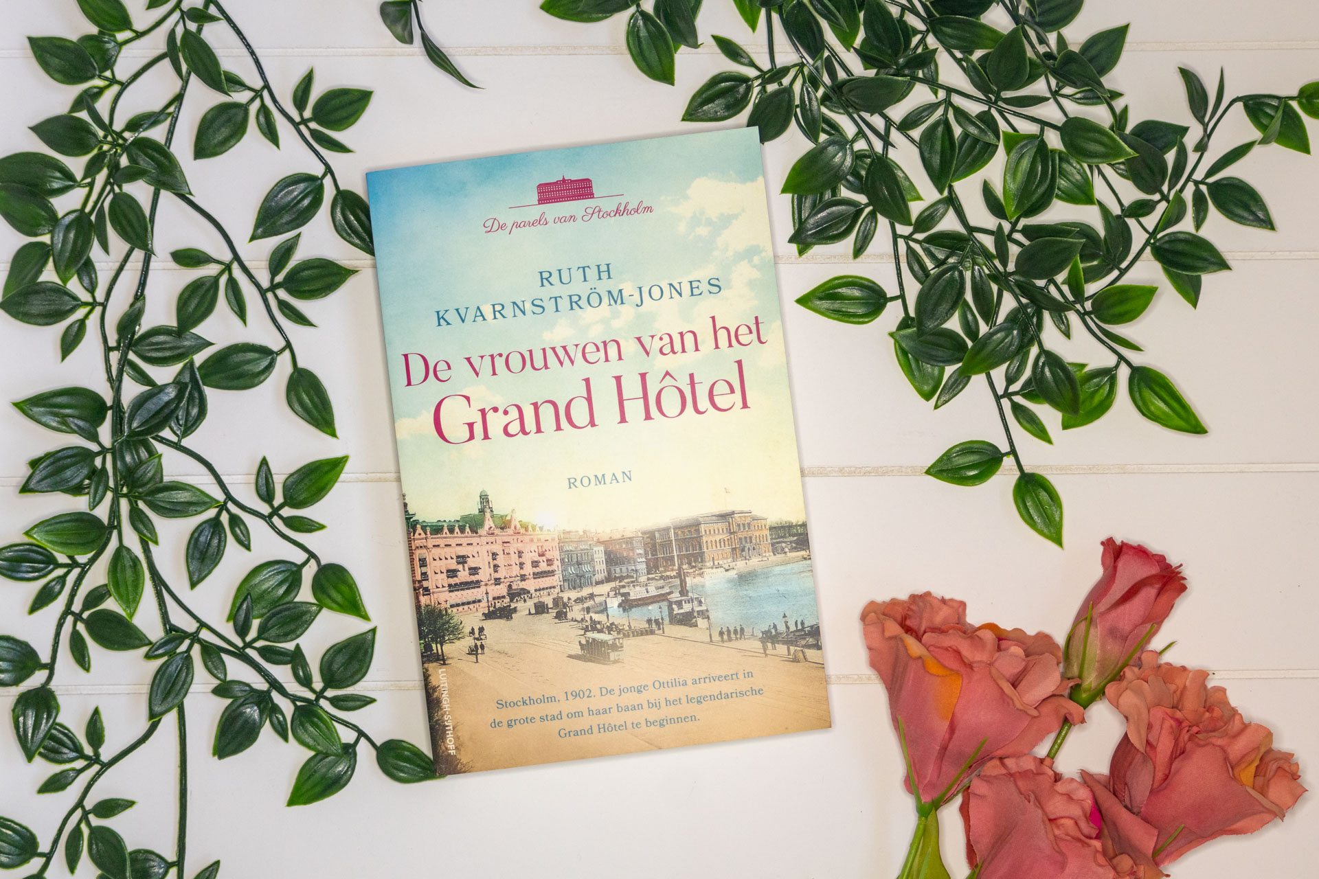 Paperback van het boek De vrouwen van het Grand Hôtel op een witte achtergrond omringd met planten en roze bloemen