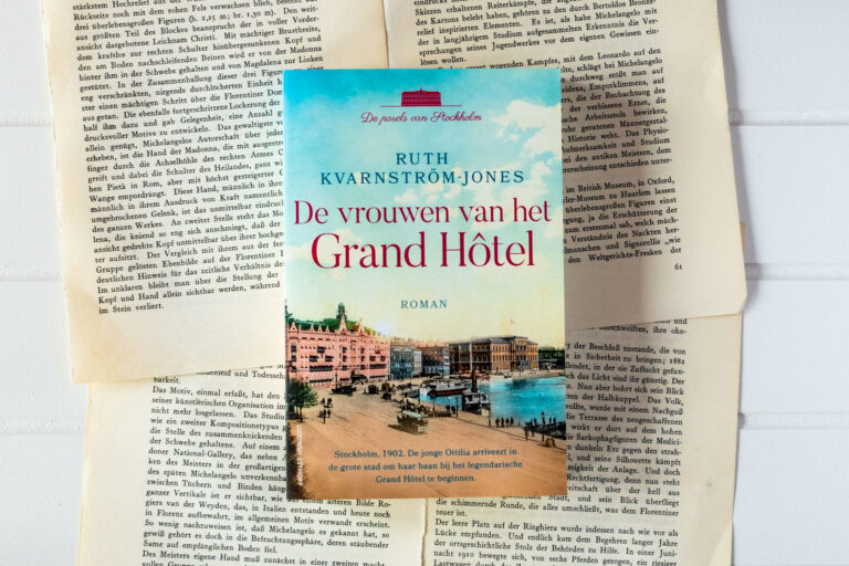 Paperback van het boek De vrouwen van het Grand Hôtel op een witte, houten achtergrond met daarop van oude, vergeelde boekpagina's.