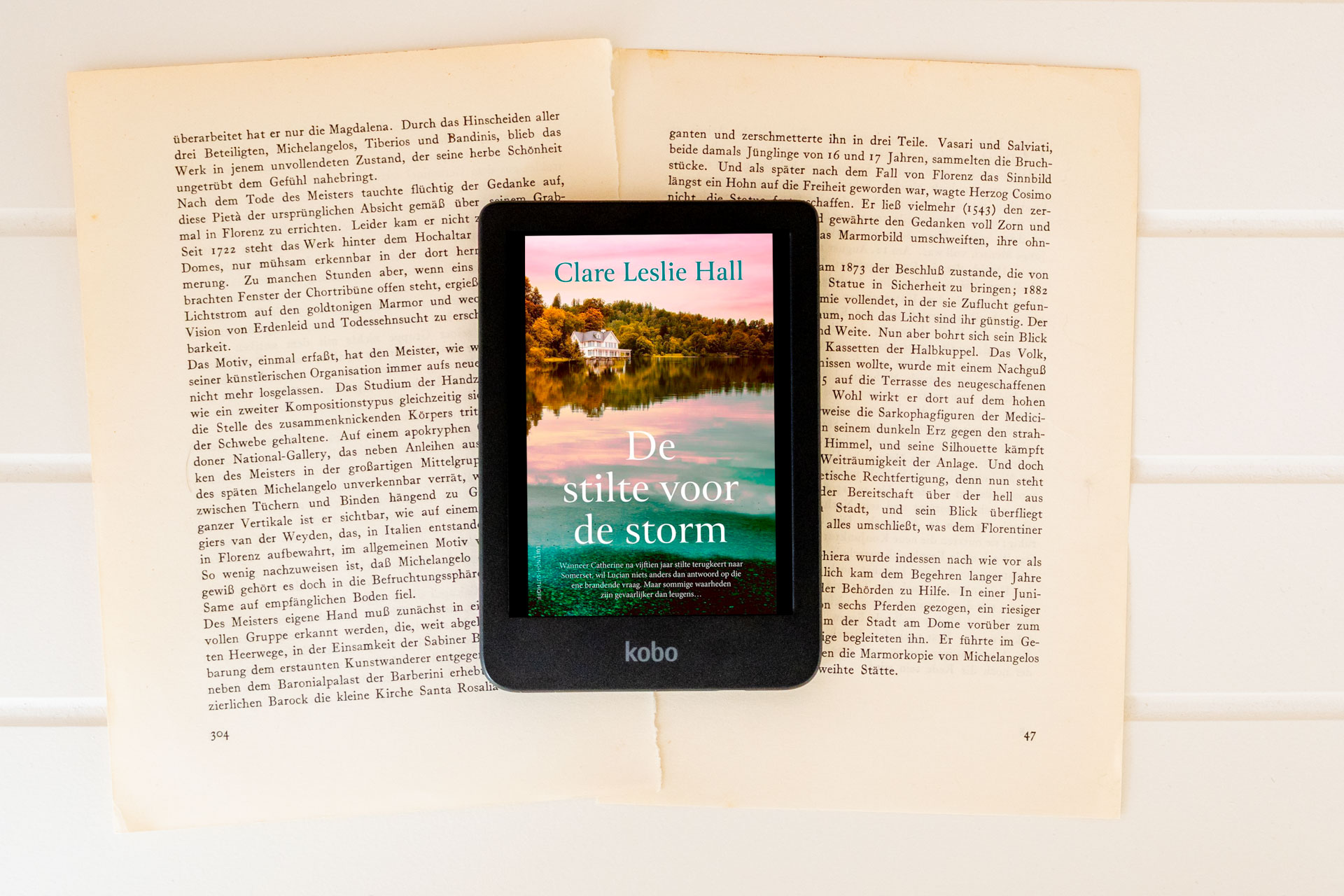 Het e-book De stilte voor de storm van Clare Leslie Hall wordt weergegeven op een e-reader die op opengeslagen, vergeelde boekpagina’s ligt. Op het scherm staat de cover met een huis aan een meer, weerspiegeld in het water, met de titel De stilte voor de storm en de naam van de auteur duidelijk zichtbaar. Onderaan de e-reader is het Kobo-logo te zien.