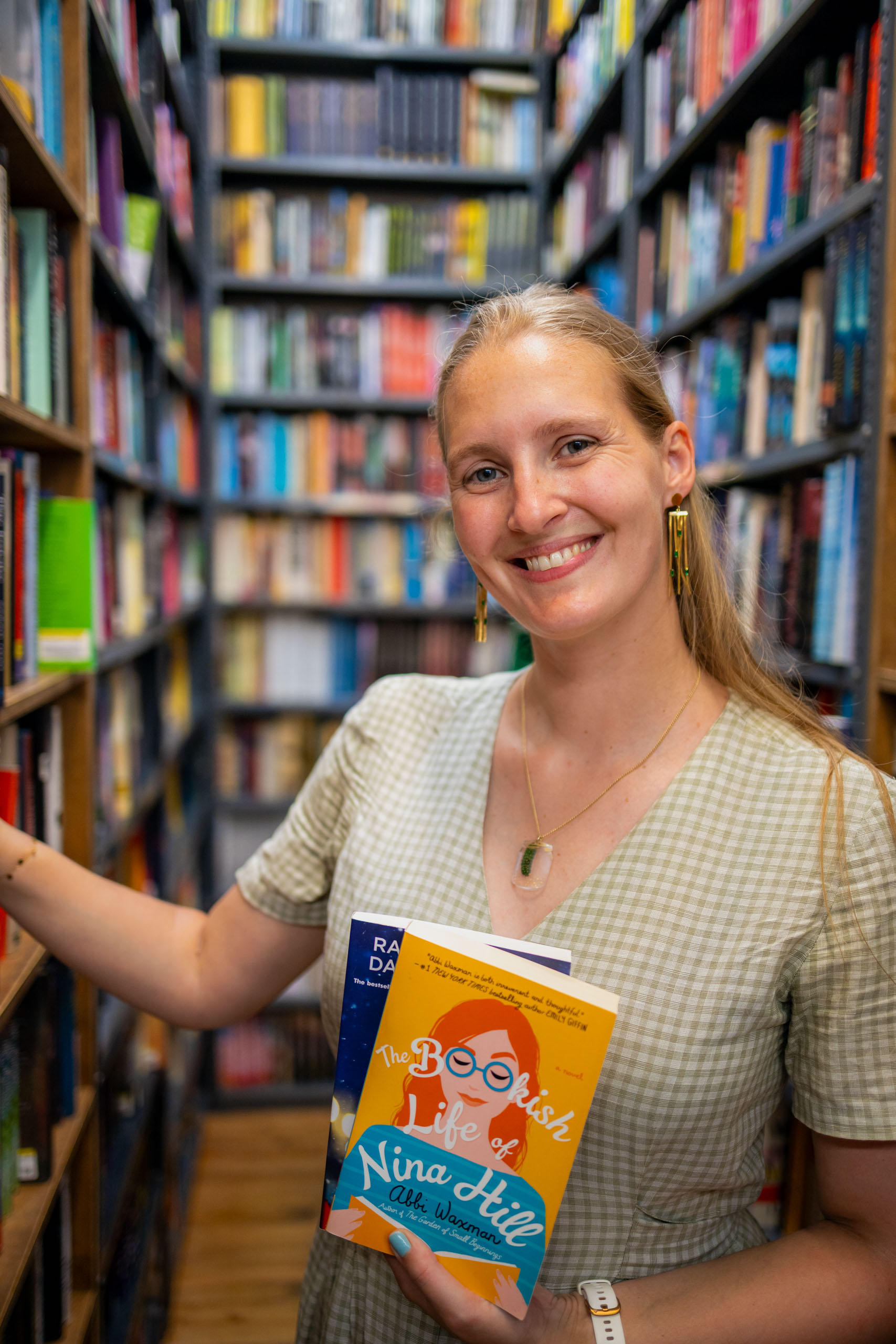 Glimlachende vrouw in een boekhandel die het boek “The Bookish Life of Nina Hill” vasthoudt, omringd door kleurrijke boekenplanken in de boekwinkel The Strand in New York. Introductiefoto voor de boekenblog Treasure in Books, waarin leesplezier en boekentips centraal staan.