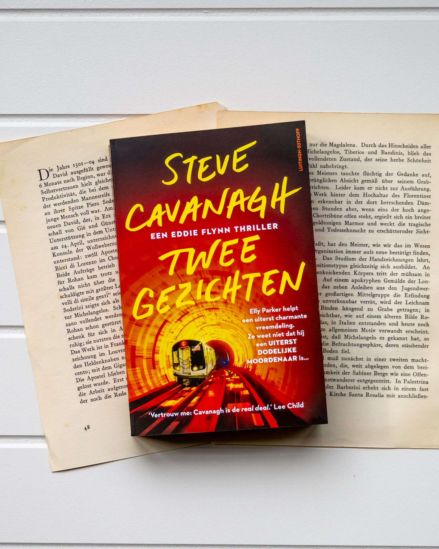Een hand houdt het boek Twee gezichten van Steve Cavanagh open boven een oude boekpagina. De cover toont een fel verlichte metrotunnel met twee treinen die op elkaar af lijken te rijden, wat een spannende en dreigende sfeer oproept.