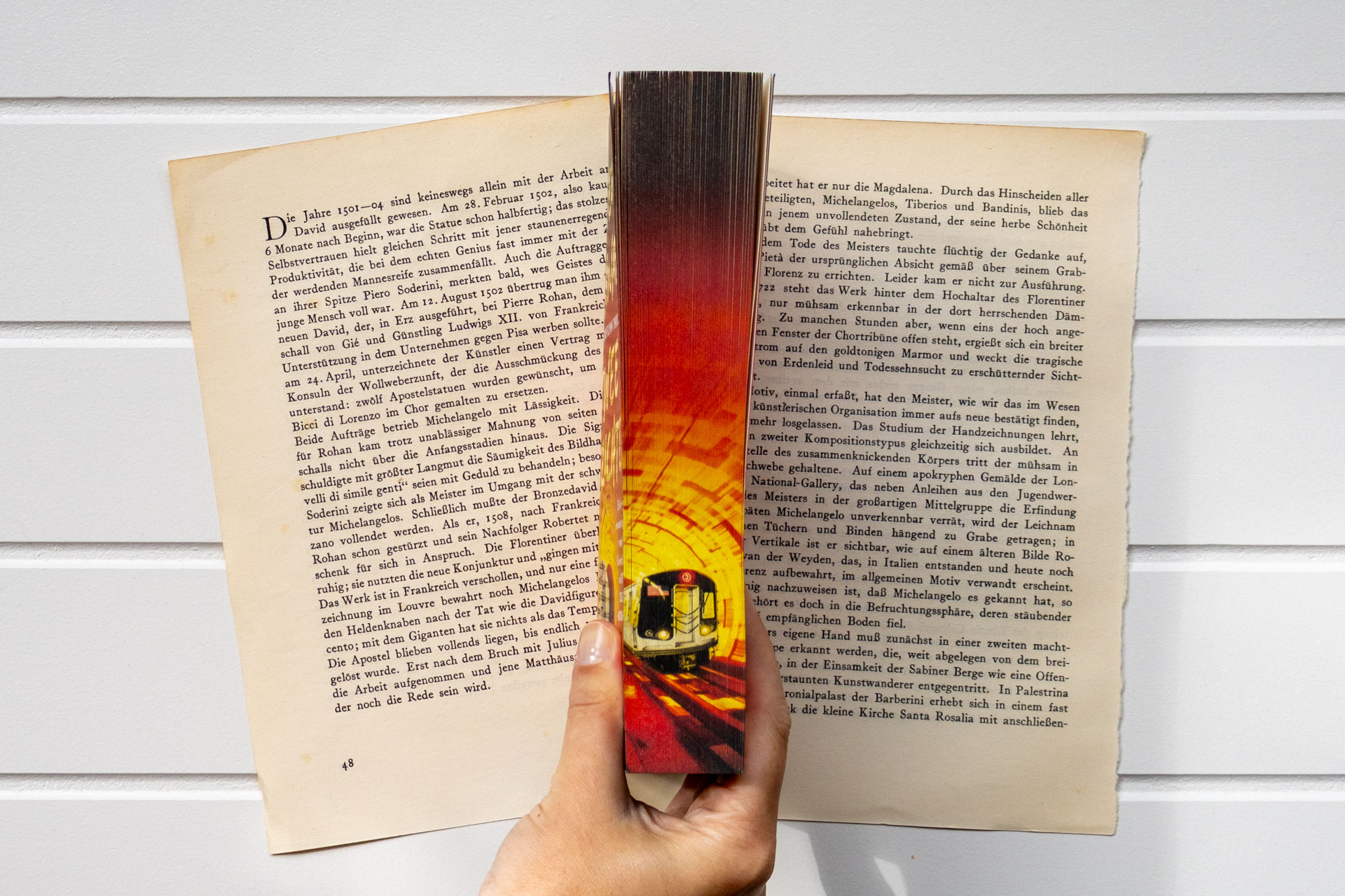 Een hand houdt het boek Twee gezichten van Steve Cavanagh open boven oude boekpagina's. Het boekblok is zichtbaar en toont een fel verlichte metrotunnel met een trein en veel verlopende kleuren rood.