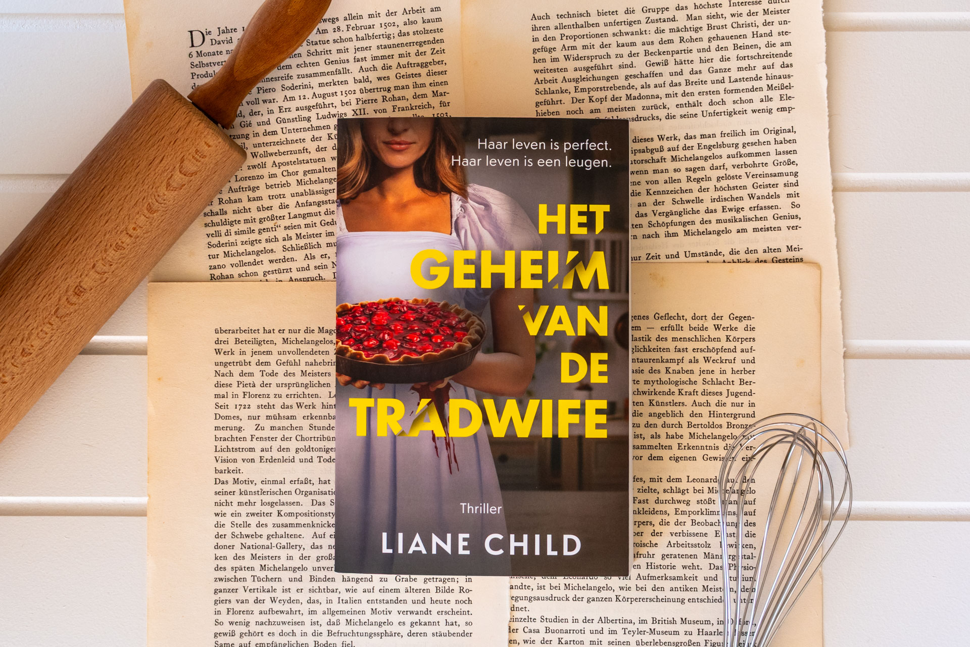 Het boek Het geheim van de Tradwife van Liane Child ligt bovenop oude boekpagina’s, met een deegroller en een garde ernaast als bakattributen. Op de cover staat een vrouw in een witte jurk met bloed erop, die een taart met dieprode vulling vasthoudt, wat een opvallend contrast creëert met haar serene uitstraling.