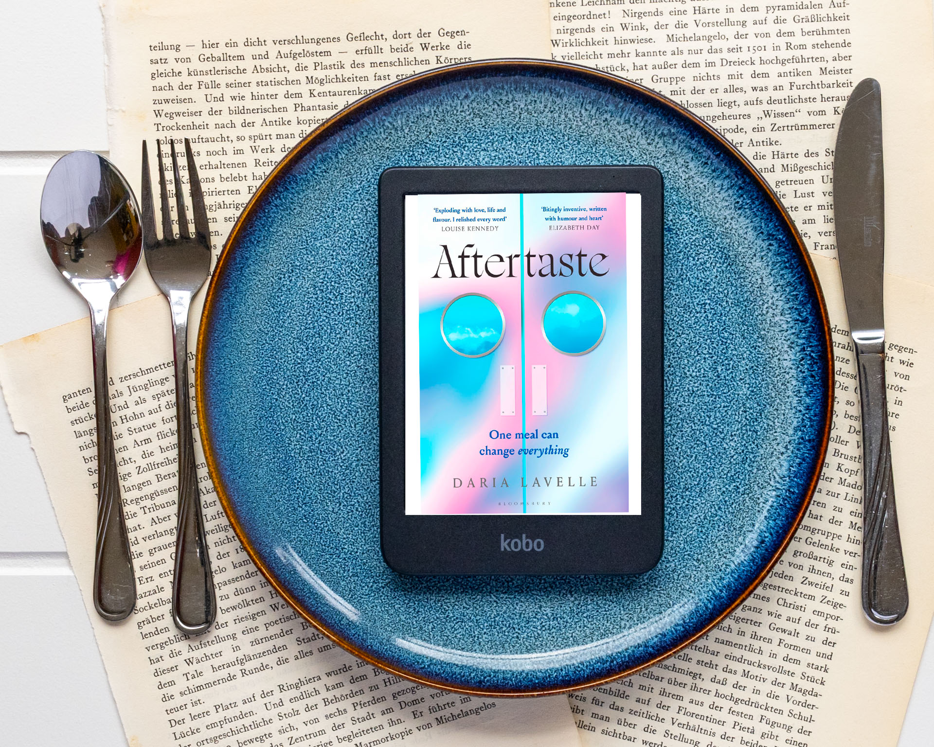Het e-book Aftertaste van Daria Lavelle wordt getoond op een e-reader die in het midden van een groot blauw bord ligt. Rond het bord liggen een vork, mes en lepel op opengeslagen, vergeelde boekpagina’s. Op het scherm is de pastelgekleurde cover zichtbaar met de titel Aftertaste, de ondertitel One meal can change everything en de naam van de auteur. Onderaan de e-reader staat het Kobo-logo.