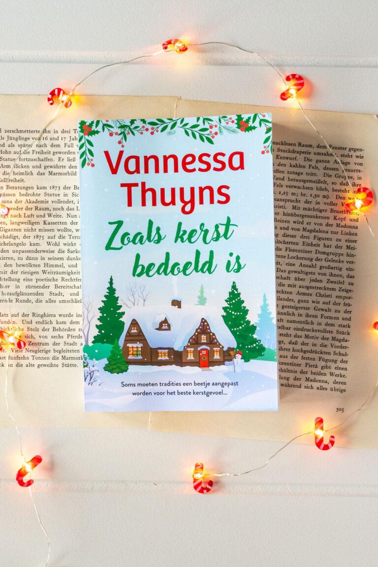 Het boek Zoals kerst bedoeld is van Vanessa Thuyns ligt op vergeelde boekpagina’s, omringd door een lichtsnoer met candy cane-lampjes. Op de cover is een sfeervol winterlandschap te zien met besneeuwde huisjes en kerstbomen.