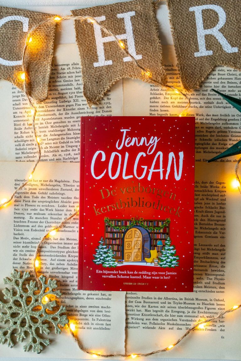 Het boek De verborgen kerstbibliotheek van Jenny Colgan ligt op opengeslagen, vergeelde boekpagina’s. Op de rode cover zijn de titel, de naam van de auteur en een illustratie van een boekenkast met een deur, boeken en twee kerstbomen te zien. Rondom het boek ligt een lichtsnoer met warmgele lampjes, een zilveren sneeuwvlokornament en bovenin hangen juten vlaggetjes met witte letters die Christmas spellen.