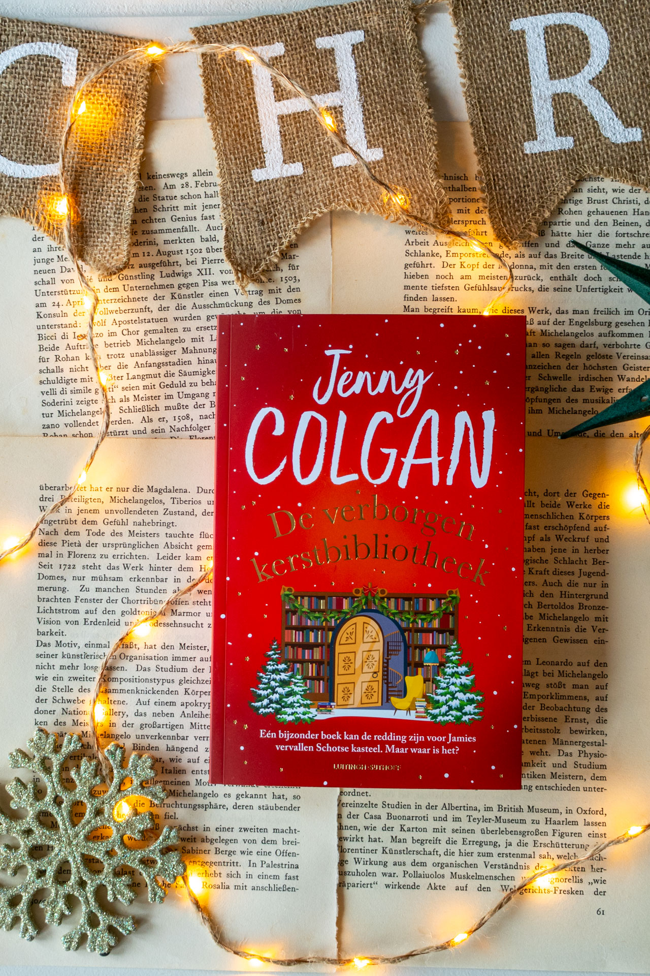 Het boek De verborgen kerstbibliotheek van Jenny Colgan ligt op opengeslagen, vergeelde boekpagina’s. Op de rode cover zijn de titel, de naam van de auteur en een illustratie van een boekenkast met een deur, boeken en twee kerstbomen te zien. Rondom het boek ligt een lichtsnoer met warmgele lampjes, een zilveren sneeuwvlokornament en bovenin hangen juten vlaggetjes met witte letters die Christmas spellen.