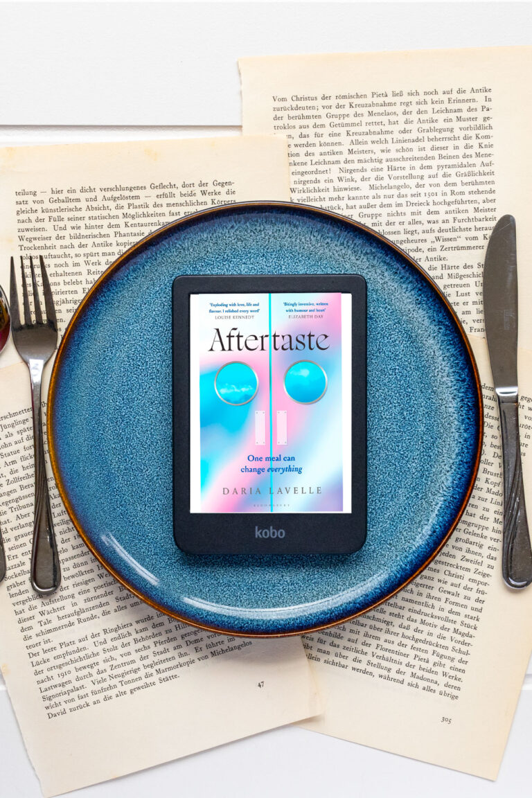 Het e-book Aftertaste van Daria Lavelle staat op een e-reader die midden op een groot blauw bord ligt. Het bord is geplaatst op opengeslagen, vergeelde boekpagina’s en wordt aan beide zijden omringd door bestek: links een vork en lepel, rechts een mes. Op het scherm is de pastelgekleurde cover zichtbaar met de titel Aftertaste, de tekst One meal can change everything en de naam van de auteur, met onderaan het Kobo-logo.