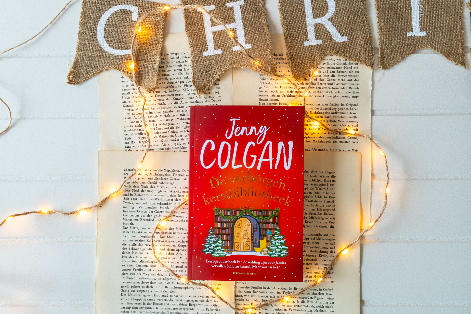 Het boek De verborgen kerstbibliotheek van Jenny Colgan ligt gecentreerd op open, vergeelde boekbladzijden. De rode omslag toont de titel en auteursnaam, met een illustratie van een boekenkast met een deur, boeken en twee kerstbomen. Langs en over de pagina’s loopt een lichtsnoer met warmgele lampjes. Bovenin is een juten vlaggetjeslijn met witte letters 'Christmas' zichtbaar.