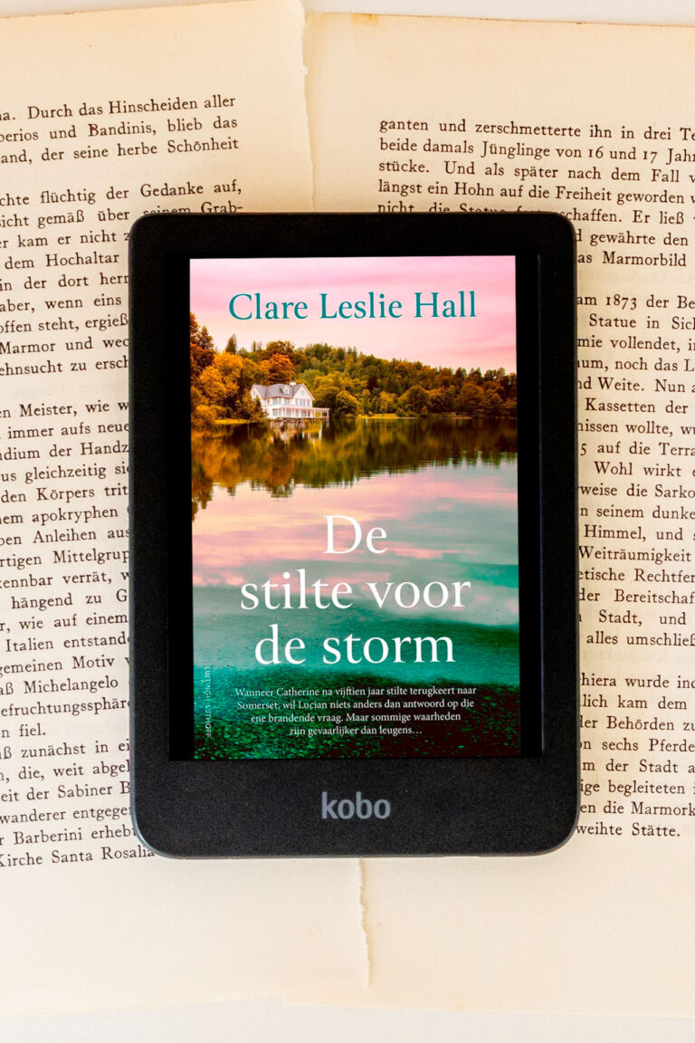 Het e-book De stilte voor de storm van Clare Leslie Hall staat afgebeeld op een e-reader die bovenop open, vergeelde boekbladen ligt. Op het scherm is een omslag te zien met een huis aan het water dat wordt weerspiegeld in het meer, met de titel De stilte voor de storm en de naam van de auteur duidelijk leesbaar. Onder aan het apparaat is het Kobo-logo zichtbaar.