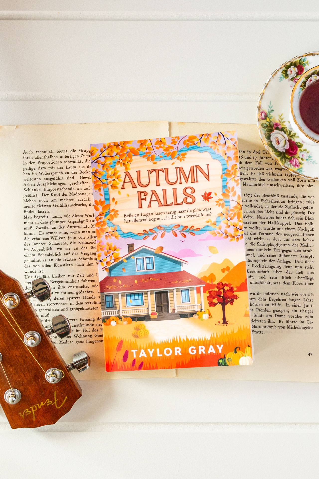 Het boek Autumn Falls van Taylor Gray ligt op opengeslagen, vergeelde pagina’s, met rechtsboven een kopje thee op een bloemig schoteltje deels in beeld. Op de cover staat een huis in een warm herfstlandschap met vallende bladeren en pompoenen, terwijl de titel Autumn Falls en de naam van de auteur duidelijk zichtbaar zijn. Linksonder is ook een deel van een gitaarhals met stemmechanieken te zien.