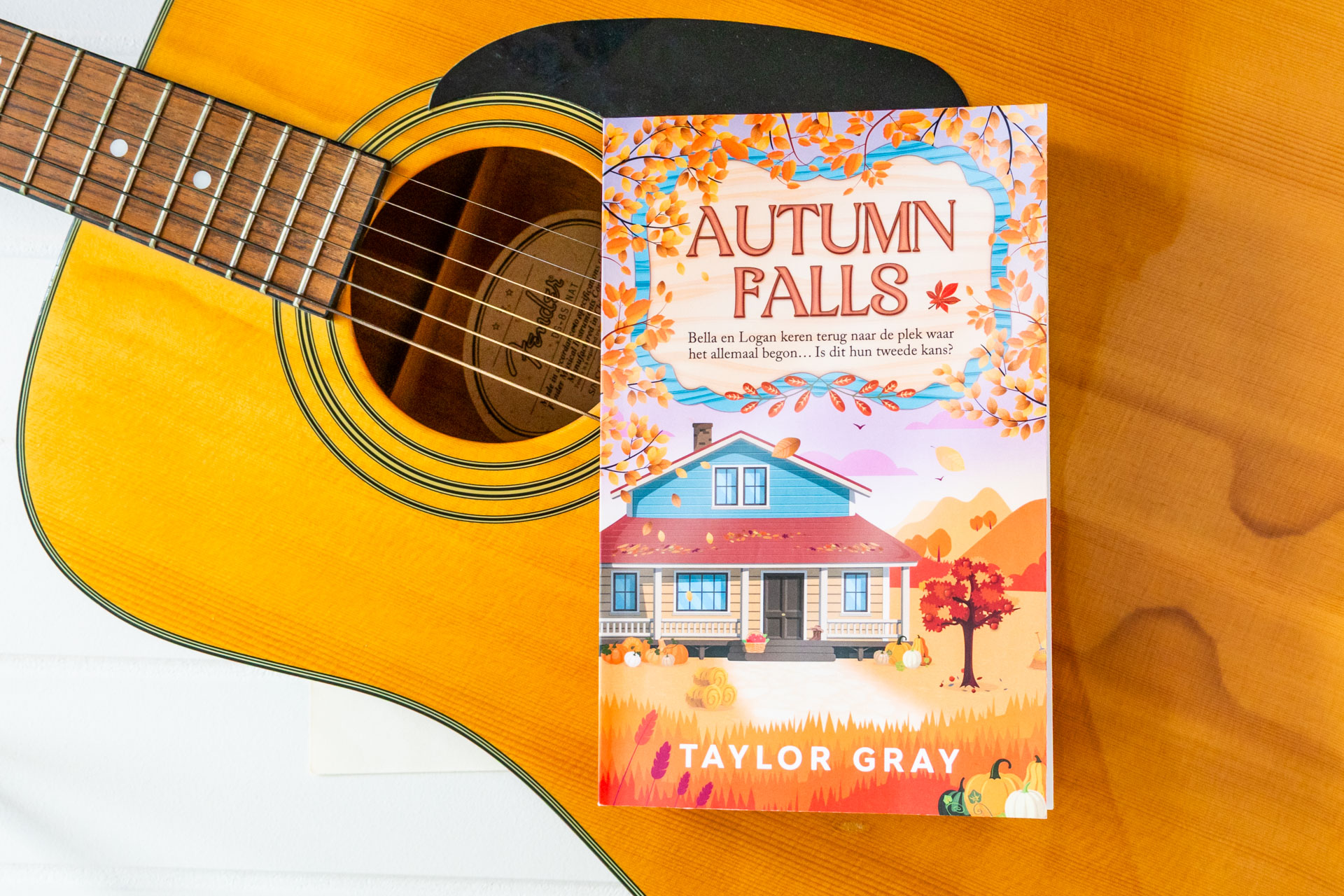 Het boek Autumn Falls van Taylor Gray ligt op de klankkast van een akoestische gitaar, waarbij de snaren en het klankgat deels zichtbaar zijn. Op de cover staat een huis in een herfstlandschap met vallende bladeren, pompoenen en warme kleuren, met de titel Autumn Falls en de naam van de auteur duidelijk in beeld.